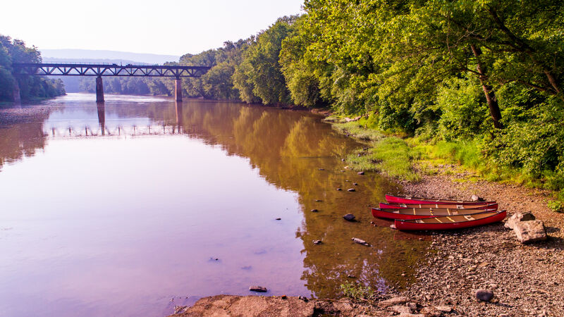 The image shows a serene river scene with a bridge in the background. The water reflects the sky and surrounding trees, creating a mirror-like effect. On the riverbank, there are three red canoes resting on the rocky shore. The lush greenery of the trees adds a vibrant contrast to the calm water and earthy tones of the bank.